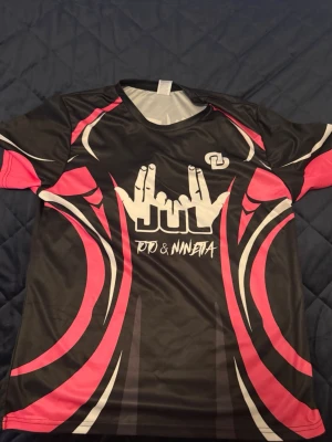 JUL T-shirt  - Cool svart sporttröja med rosa och vita detaljer, tryckta händer och texten JUL på framsidan samt siffran 13 och namnet Ninneta på ryggen. Tillverkad i ett lätt och snabbtorkande material, perfekt för träning eller lagidrott.