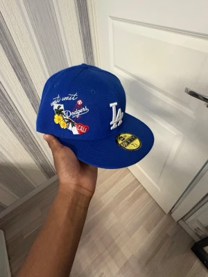 Blå LA Dodgers keps från New Era - Snygg blå keps från New Era med LA Dodgers-logga framtill och broderade detaljer på sidan som palm, text och Cali-märke. Kepsen har platt skärm och klassisk snapback-stil. Perfekt för dig som vill sticka ut med sportig vibe.
