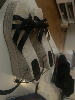 Beiga espadrilles med svarta band - Säljer ett par snygga espadrilles med hög platåsula i flätad jute och breda svarta band som korsas över foten. Sandalerna har öppen tå och justerbar rem runt ankeln. Perfekta för sommaren och ger en trendig look till din outfit. Oanvända