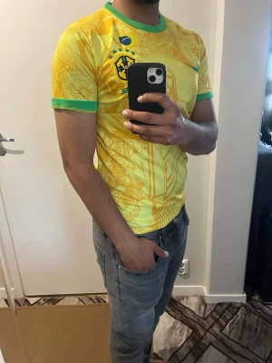 Gul Brasilien fotbollströja Nike - Snygg gul Brasilien fotbollströja från Nike med gröna detaljer vid krage och ärmslut. Tröjan har ett stort motiv av Kristusstatyn och brasilianska symboler, samt Brasilien-logga och Nike-logga på bröstet. Perfekt för dig som älskar fotboll och vill sticka ut.