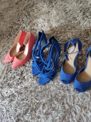 Två par pumps i rosa och blått - Säljer två par snygga pumps, ett par i korallrosa med spetsig tå och ett par i klarblått med öppen tå och remmar runt ankeln. Båda paren har hög klack och är i mockaimitation. Perfekta för dig som vill sticka ut med färg på fötterna.