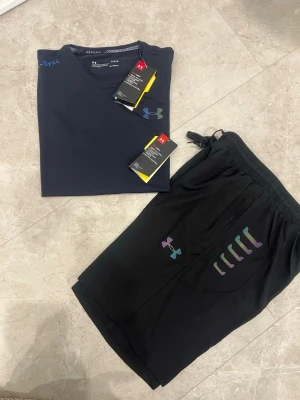 Under Armour träningsset t-shirt & shorts - Snyggt träningsset från Under Armour med en mörkblå t-shirt och svarta shorts. T-shirten har logga på bröstet och är tillverkad i ett snabbtorkande funktionsmaterial. Shortsen har elastisk midja, dragsko och färgglada detaljer samt logga på benet.
