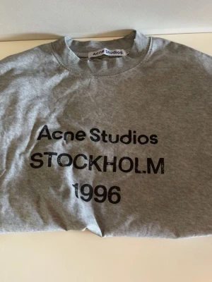 Acne Studios long sleeve - Snygg grå långärmad topp från Acne Studios med tryck 'STOCKHOLM 1996' framtill. Klassisk rund halsringning och relaxed fit. Perfekt till jeans eller kjol för en clean och trendig look.