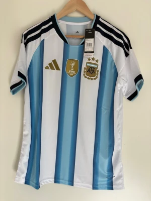 Argentina matchtröja Adidas M - Säljer en officiell Argentina fotbollströja från Adidas i storlek M. Tröjan har blå och vita ränder, svarta detaljer på axlarna, guldiga loggor och emblem samt AFA-märke med tre stjärnor. Slim fit-modell i lätt och ventilerande material.