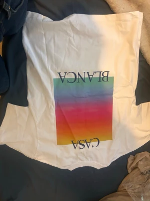 Vit Casa Blanca t-shirt med färgglad print - Säljer en vit t-shirt från Casa Blanca med en stor färgglad rektangel i regnbågens färger och texten 'CASA BLANCA' på ryggen samt en mindre likadan print på bröstet. Klassisk passform och rund hals. Perfekt för dig som gillar stilrena men ändå färgstarka plagg.