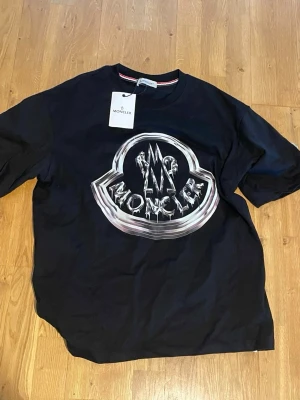 Svart Moncler t-shirt med stor logga - Snygg svart t-shirt från Moncler med stor, glansig logga på bröstet och ett mindre Moncler-märke på ärmen. Klassisk rund hals och korta ärmar. Perfekt för dig som gillar streetwear och vill sticka ut med ett exklusivt plagg.