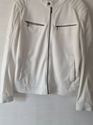 Vit bikerinspirerad jacka från zara  - Snygg vit jacka med bikerstil, raka linjer och ståkrage. Jackan har två bröstfickor med dragkedja och två sidofickor. Dekorativa sömmar på axlar och rygg ger extra edge. Materialet ser ut att vara syntetiskt, troligen polyester.