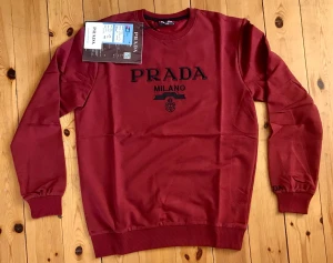 Vinröd sweatshirt från Prada - Snygg vinröd sweatshirt från Prada med svart broderad logga och texten 'Milano' på bröstet. Tröjan har rund halsringning, ribbade muddar vid ärmslut och nederkant samt en klassisk passform. Perfekt för en trendig och avslappnad stil.
