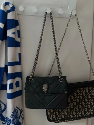 Svart quiltad handväska från Kurt Geiger - Snygg svart quiltad handväska från Kurt Geiger med silverfärgad kedjerem och ikoniskt örnhuvud i metall framtill. Väskan är tillverkad i läder och har en stilren, rektangulär form som ger en lyxig känsla.