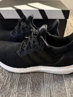 Adidas Ultra Boost svarta sneakers - Svarta Adidas Ultra Boost sneakers med stickad ovandel och klassiska tre ränder på sidorna. Skorna har en vit Boost-sula för extra komfort och svart hälkappa. Perfekta för dig som gillar sportig och stilren look. (Storlek 44)