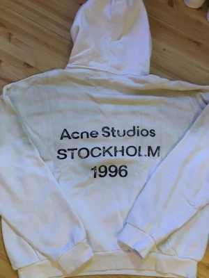 Vit hoodie Acne Studios Stockholm - Vit hoodie från Acne Studios med tryck på ryggen där det står 'Acne Studios STOCKHOLM 1996'. Klassisk huva och ribbade muddar vid ärmslut och nederkant. Mjuk insida och avslappnad passform, perfekt för en cool och enkel streetwear-look.