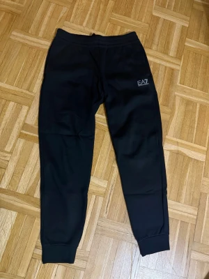 Emporio Armani EA7 Byxor - Svarta mjukisbyxor från EA7 med logga på vänster lår. Byxorna har resår i midjan och vid bensluten för en skön passform. Perfekta för en avslappnad stil och tillverkade i mjukt material. Helt nya har aldrig använts. Storlek S. Köpta från JD för 1499kr
