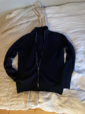 Mörkblå stickad ziptröja - Mörkblå stickad tröja med dragkedja framtill och hög krage. Tröjan har lång ärm och ribbad struktur, perfekt för lager på lager. Enkel och stilren design utan synligt märke.