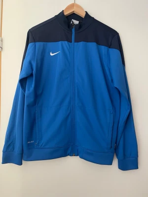 ” Barn” Blå Nike Dri-FIT träningsjacka - Snygg blå träningsjacka från Nike med svart axelparti och vit logga på bröstet. Jackan har hel dragkedja, ståkrage och är tillverkad i Dri-FIT-material som transporterar bort fukt. Perfekt för träning eller uppvärmning.