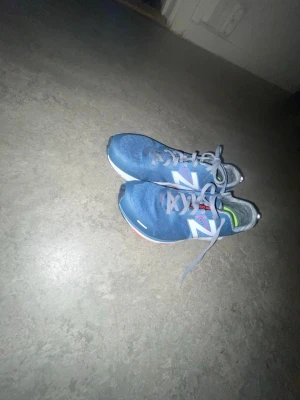 Blå New Balance sneakers - Snygga blå sneakers från New Balance med vita och grå detaljer samt den klassiska N-loggan på sidan. Skorna har meshmaterial för bra andningsförmåga och grå skosnören. Sulan har inslag av vitt och rött, och insidan har gröna detaljer.