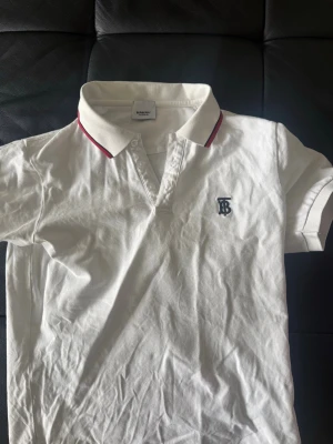 Vit pikétröja från Burberry - Klassisk vit pikétröja från Burberry med korta ärmar och krage med röd detalj. Tröjan har Burberrys ikoniska TB-logga broderad på bröstet och stängs med knappar framtill. Perfekt till både jeans och kjol. L men mer som M