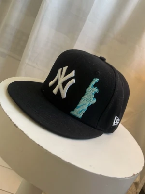 New era keps med brodyr - Cool svart keps från New Era med NY Yankees-logga i vitt och broderad Frihetsgudinna i blått på framsidan. Baksidan har ett broderat rött äpple med grönt blad och MLB-logga. Klassisk flat peak och snapback-modell, perfekt för dig som gillar streetstyle. Storleken är 56,8