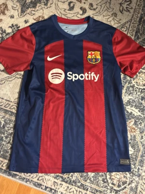 Barcelona matchtröja Nike blå/röd - Säljer en FC Barcelona matchtröja från Nike med klassiska blå och röda vertikala ränder. Tröjan har klubbens emblem, Nike-logga och Spotify som sponsor på bröstet. Materialet är lätt och andas, perfekt för fotboll eller träning.