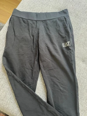Svarta mjukisbyxor EA7 Emporio Armani - Svarta mjukisbyxor från EA7 Emporio Armani med logotyp på vänster ben. Byxorna har elastisk midja och sidofickor, tillverkade i mjukt bomullsmaterial. Perfekta för en chill och avslappnad look.