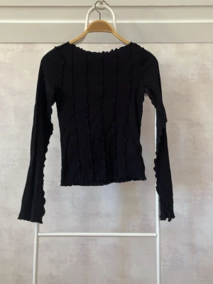 Svart långärmad topp med volangdetaljer - Trendig svart långärmad topp med vågiga volangdetaljer längs ärmar, kropp och kanter. Figurnära passform och stretchigt material som ger en snygg siluett. Perfekt att styla med jeans eller kjol för en cool look.