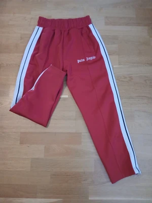 Röda mjukisbyxor Palm Angels - Säljer ett par röda mjukisbyxor från Palm Angels med vita sidoränder och logga tryckt på vänster sida. Byxorna har resår i midjan och raka ben. Perfekta för en avslappnad och trendig streetwear-look.