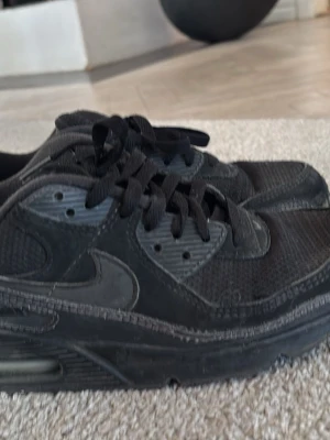 Svarta Nike Air Max sneakers - Säljer ett par helsvarta Nike Air Max sneakers med klassisk siluett. Skorna har mesh och syntetmaterial i ovandelen, samt den ikoniska Air Max-sulan för extra komfort. Swoosh-logga på sidan och svarta skosnören. Perfekta för en streetwear-look.