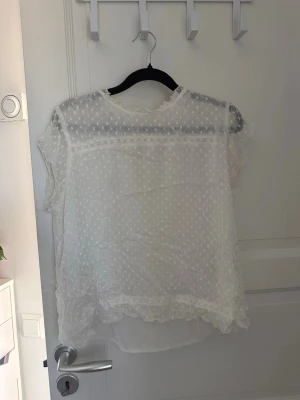 Vit transparent blus med spetsdetaljer - Söt vit blus i transparent material med små prickar och spetsdetaljer vid hals, ärmslut och nederkant. Blusen har rund halsringning och volangkantade korta ärmar. Perfekt för en romantisk och luftig look.