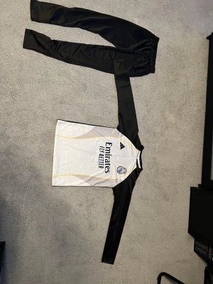Real Madrid träningsset från Adidas - Säljer ett Real Madrid träningsset från Adidas med långärmad tröja och matchande svarta träningsbyxor. Tröjan är vit med svarta ärmar, gula detaljer och klubbens logga samt Emirates Fly Better-tryck. Materialet är lätt och andas, perfekt för fotbollsträning.