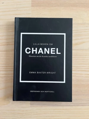 Lilla boken om Chanel - En svart, hård inbunden bok med vit text och ram, skriven av Emma Baxter-Wright. Boken berättar historien om det ikoniska modehuset Chanel och Coco Chanels liv och inflytande inom modevärlden. Perfekt för den modeintresserade och nyfikna läsaren.