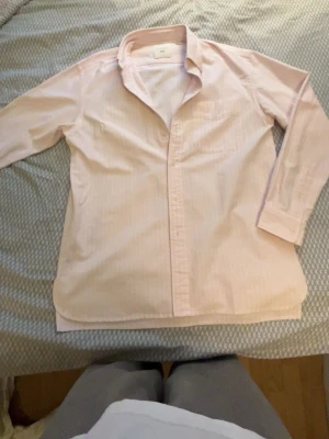 Ljusrosa skjorta från JW Anderson x Uniqlo - Ljusrosa skjorta med diskreta vita ränder från JW Anderson x Uniqlo. Klassisk krage, långärmad och bröstficka. Skjortan är gjord i mjukt bomullsmaterial och har en avslappnad passform. Perfekt för dig som gillar stilrena plagg. Skjortan har en liten fläck men den märks knappt