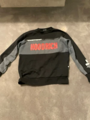 Svart och grå HOODRICH sweatshirt - Cool svart och grå sweatshirt från Hoodrich med stor röd logga på bröstet och tryck på ena ärmen. Tröjan har rund hals, ribbade muddar och kontrastpaneler på ärmarna. Perfekt för en streetwear-look.