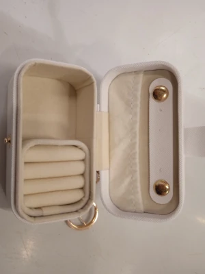 Vit liten smyckesväska med gulddetaljer - Snygg och kompakt vit smyckesväska med guldfärgade detaljer och nyckelring. Väskan har mjukt foder och flera fack för ringar och småsaker. Perfekt att hänga på väskan eller använda som förvaring för smycken när du är på språng.