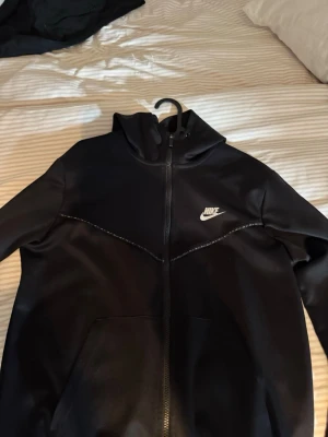 Svart Nike hoodie med dragkedja - Svart hoodie från Nike med vit logga på bröstet och hel dragkedja framtill. Hoodien har en huva och två fickor på framsidan. Materialet är mjukt och sportigt, perfekt för chill eller träning. Snygg design med diskreta sömmar över bröstet.