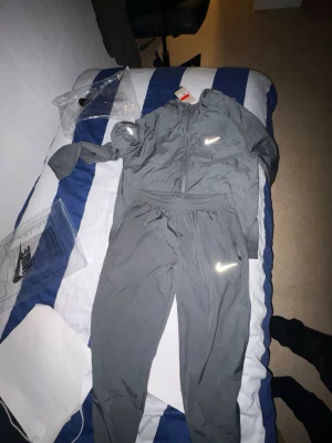 Nike Miller  - Säljer ett grått träningsset från Nike med jacka och byxor. Setet har reflekterande Nike-loggor på både jackan och byxorna. Materialet är lätt och passar perfekt för löpning eller gym. Jackan har dragkedja och elastiska muddar. Perfekt för dig som vill ha stil och funktion.