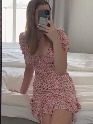 Rosa blommig klänning - Söt kort klänning med rosa blommönster och puffiga korta ärmar. Klänningen har v-ringning och volangdetaljer längs nederkanten, även knytband i sidan som detalj. Något liten vid brösten. Perfekt för sommaren