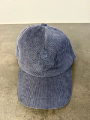 Blå manchesterkeps från Loro Piana - Säljer en snygg blå keps i manchestertyg från Loro Piana. Kepsen har böjd skärm, broderad logga framtill och justerbart spänne bak. Perfekt accessoar för en avslappnad och trendig stil.
