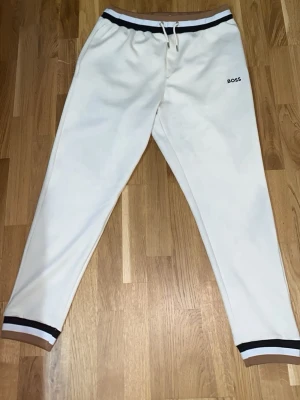 Vita mjukisbyxor från BOSS - Snygga vita mjukisbyxor från BOSS med svart logotyp på låret. Byxorna har svarta detaljer vid midja och benslut samt dragsko i midjan. Perfekta för chill eller sportiga dagar, tillverkade i mjukt material som känns skönt mot huden.