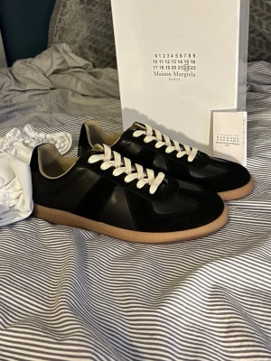 Maison Margiela svarta sneakers - Maison Margiela sneakers i svart läder och mocka med vita snören och beige innersula. Klassisk låg modell med gummisula i naturfärg. Snygg och stilren design som passar till det mesta. Kommer med originalkartong och dustbag.0bs; lnte 0rglnal, men mycket bra kvalitet. Skorna är gjorda i äkta skinn 