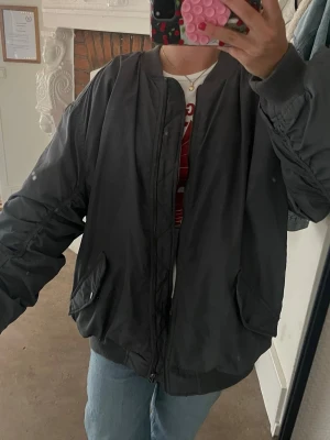 Oversized svart bomberjacka - Snygg oversized svart bomberjacka med dragkedja framtill och två stora fickor. Jackan har ribbade muddar vid ärmslut och midja samt en cool rynkad detalj längs ryggen. Perfekt för en streetwear-look och enkel att matcha med jeans eller hoodie.