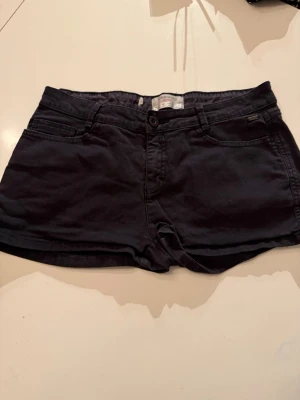 Svarta shorts från Springfield - Säljer ett par svarta shorts från Springfield Essentials. De har klassisk femficksdesign, knapp och dragkedja framtill samt låg midja. Materialet är mjukt och känns som bomull, perfekt för varma dagar. Enkla att matcha med olika toppar.