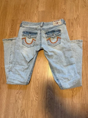 True Religion ljusa bootcut jeans - Säljer ett par ljusa blå bootcut jeans från True Religion med ikoniska broderade bakfickor och silverfärgade knappar. Jeansen har klassisk passform och är tillverkade i denim med snygga detaljer på fickorna. I storlek W25 som jag tror är typ M eller S