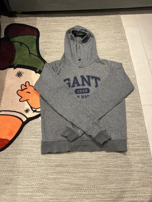 Gant hoodie storlek 158/164 cm  - Säljer en grå hoodie från GANT med blått tryck på bröstet och texten '1949 New Haven'. Tröjan har huva, ribbade muddar och är tillverkad i mjukt material som passar perfekt för en avslappnad look.