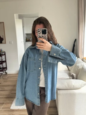 Oversize blå jeansjacka - Säljer en oversize blå jeansjacka med klassisk krage och stora bröstfickor. Jackan har knappar framtill och på ärmsluten, samt raka ärmar och en avslappnad passform. Perfekt att slänga över en t-shirt för en cool look.