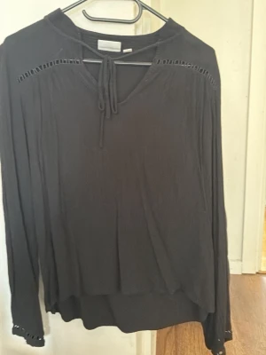 Svart långärmad blus från Vila - Svart blus från Vila med långa ärmar och dekorativa håldetaljer vid axlarna. Blusen har en v-ringning med knytband och är tillverkad i ett lätt och luftigt material som faller snyggt. Perfekt för att skapa en stilren look.