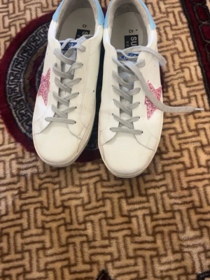 Vita sneakers med rosa glitterstjärna - Snygga vita sneakers från Superstar med gråa skosnören och en rosa glitterstjärna på sidan. Skorna har ljusblå detalj på hälen och är tillverkade i läder. Perfekt för dig som vill sticka ut med coola detaljer och klassisk design.