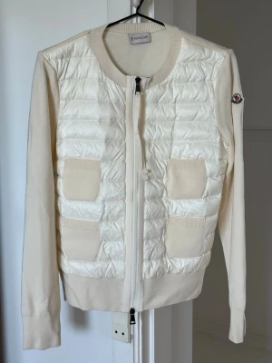 Vit cardiganjacka från Moncler - Snygg vit cardiganjacka från Moncler med quiltad front och stickade ärmar. Jackan har två ribbade fickor fram, rund halsringning och dragkedja. Moncler-logga på ärmen. Perfekt för lager-på-lager och stilrena outfits.