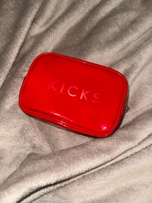 Röd necessär från Kicks med dragkedja - Snygg röd necessär från Kicks i blankt syntetmaterial. Väskan har en praktisk dragkedja och en stilren, rektangulär form. Perfekt för att organisera smink eller småprylar i väskan. Kicks-loggan syns tydligt på framsidan.