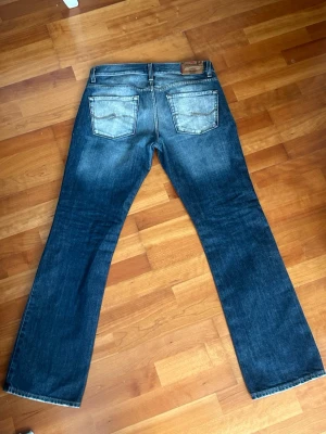 Blå vintage jeans från Jack and Jones - Säljer ett par klassiska blå bootcut jeans från Jack & Jones med slitningar och snygg tvätt. Jeansen har fem fickor, normal midja och är tillverkade i denim. Perfekta för dig som gillar en avslappnad men ändå stilren look.