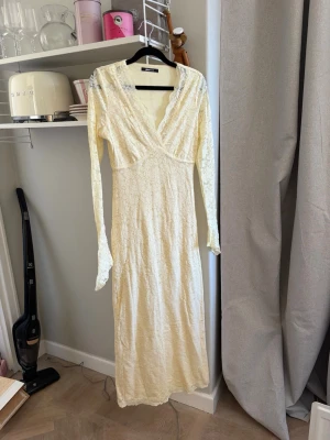 Lång spetsklänning från Gina Tricot - Elegant långklänning i ljus cremefärg från Gina Tricot. Klänningen har omlott-ringning, långa ärmar och är helt täckt av vacker spets. Perfekt för dig som vill ha en romantisk och feminin look. I storlek S💕