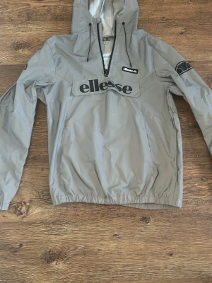  Grå vindjacka från Ellesse med huva - Snygg grå vindjacka från Ellesse med huva och dragkedja framtill. Jackan har logotyp på bröstet och ärmen, elastisk nederkant och långa ärmar. Materialet är lätt och passar perfekt för blåsiga dagar.
